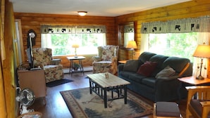 A cozy cabin-style room with a green sofa, two armchairs, a wooden coffee table, and a small side table. There are two windows with checkered curtains, and a lamp on each side table.