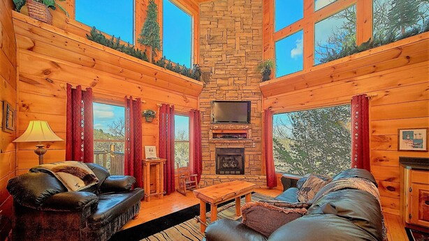 A cozy cabin interior with a stone fireplace, leather sofas, and large windows offering a view of the surrounding forest. - Aug Special-Secluded, Privacy, Mountain View, Sunset, 3D Movies, Hot Tub,Game Rm (Sevierville)