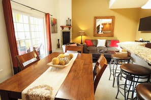 A dining area with a wooden table, chairs, a tray of food, a sofa, and a bar stool. - Leavenworth Vacation Rental, Luxury , Downtown, Amazing Mountain View Condo, (Leavenworth)