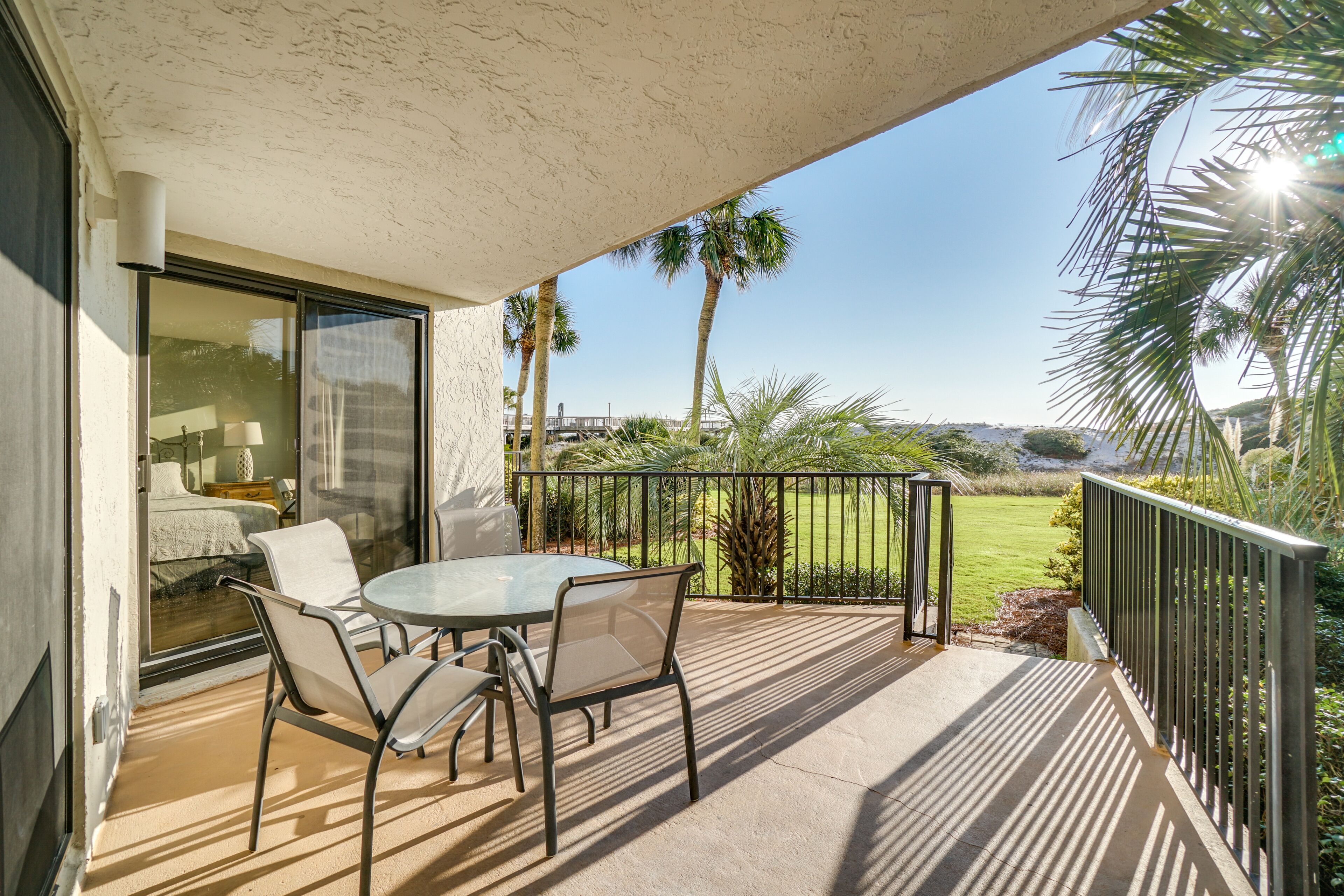 A balcony with a glass door leading to a bedroom, a round table with chairs, and a view of a lawn and palm trees.