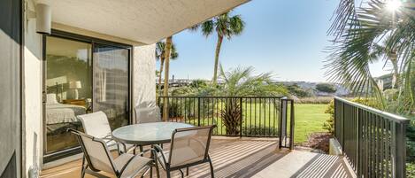 A balcony with a glass door leading to a bedroom, a round table with chairs, and a view of a lawn and palm trees.