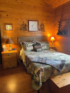 A wooden cabin-style bedroom with a bed, two bedside tables, a nightstand, a wall-mounted clock, framed artwork, and a decorative wall piece. - Private Lakefront Cabin with Dock. Fish, Boat, Relax & Enjoy Romantic Sunsets!!! (Fort Gaines)