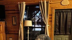 Ein gemütliches Cabin-Interieur mit Holzboden, Schaukelstuhl, Lampe und einem Fenster mit Vorhängen.