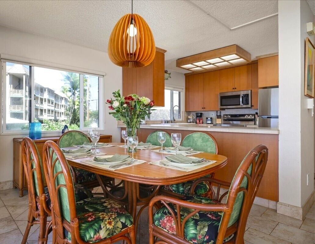 Comedor con mesa de madera dispuesta para seis personas, rodeada de sillas de mimbre con tapicería floral. Un ventanal ofrece vistas a un edificio con balcones.