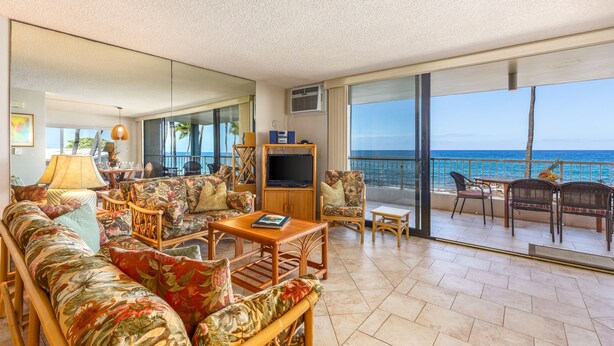 A spacious living room with a large sofa, a coffee table, and a TV. There is a balcony with a view of the ocean.