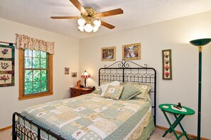 A bedroom with a bed, a nightstand, a lamp, a green side table, a ceiling fan, and a window with blinds.