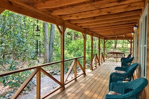 Een overdekt houten terras met een balustrade, omringd door weelderige begroeiing en bomen.