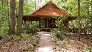 A wooden cabin with a covered porch and a stone pathway leading to it, surrounded by trees.