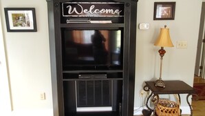 A fireplace with a 'Welcome' sign above it, a lamp on a side table, and framed pictures on the wall. - Hawk's Nest Hideaway With Creek That Runs Through This Very Private Property (Mentone)