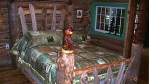 Un dormitorio rústico de cabaña de troncos con una cama de madera, una figurita de oso y una ventana con vistas a los árboles.
