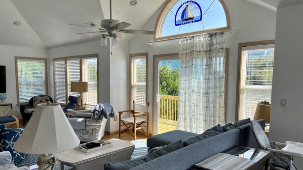 Un salon spacieux avec un grand canapé, une table basse et un ventilateur de plafond. Il y a plusieurs fenêtres équipées de stores, un balcon avec vue sur la verdure et une fenêtre décorative en forme de voilier.