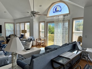 A spacious living room with a large sofa, a coffee table, and a ceiling fan. There are multiple windows with blinds, a balcony with a view of greenery, and a decorative sailboat window.