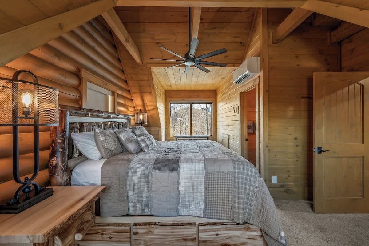 Un dormitorio tipo cabaña de troncos con cama, ventilador de techo, mesita de noche con lámpara y puerta de madera.