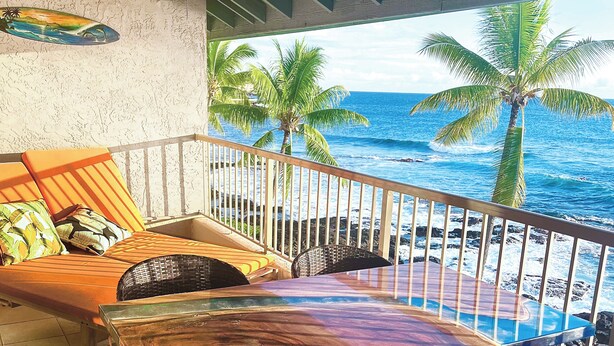 A balcony with a view of the ocean, featuring a wooden bench and a cushioned seating area with pillows.