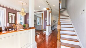 Un pasillo espacioso con una escalera de madera, un candelabro y vistas a un salón.