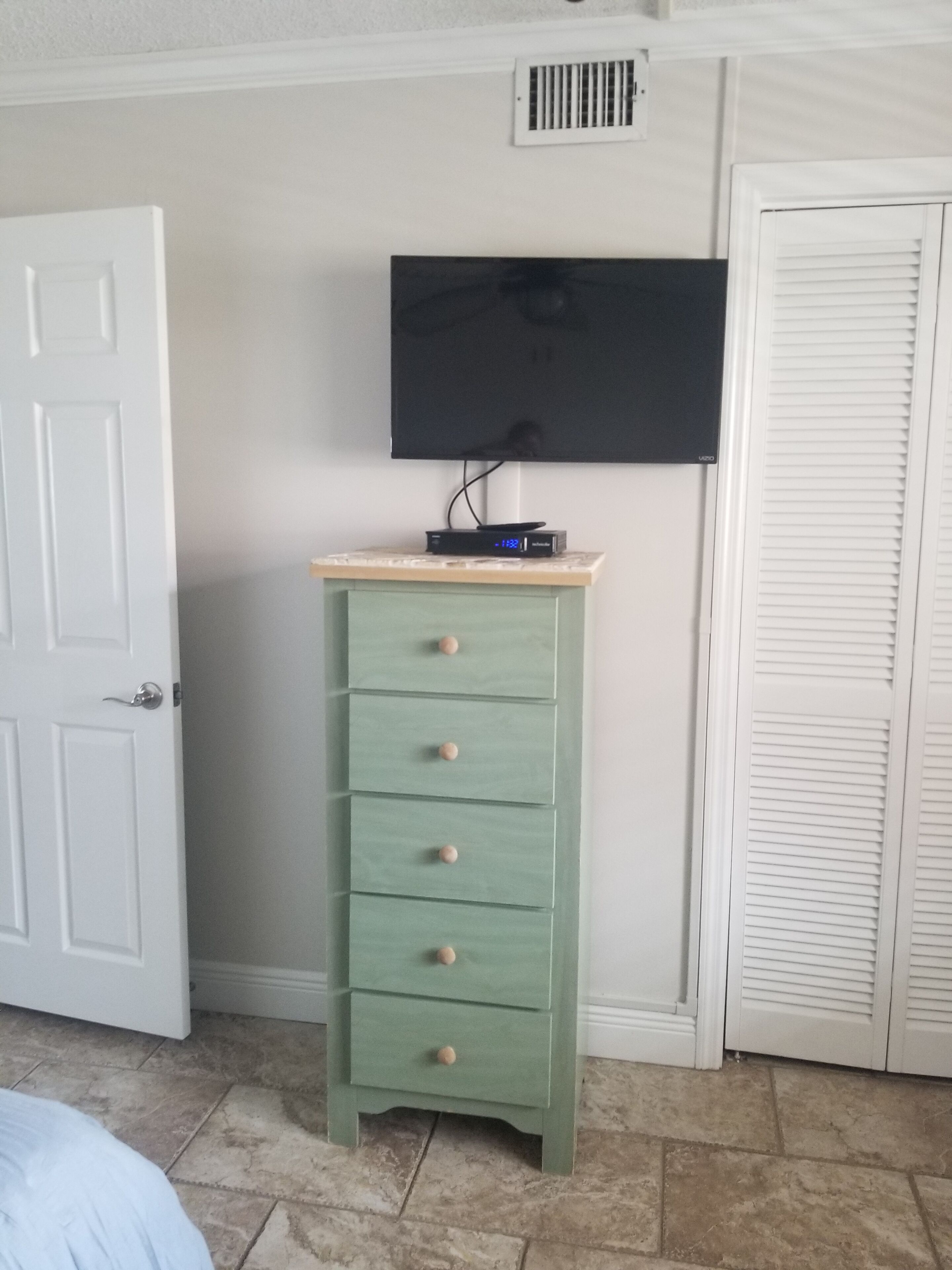 A room with a white door, a green chest of drawers, a flat-screen TV mounted on the wall, and a white closet with louvered doors.