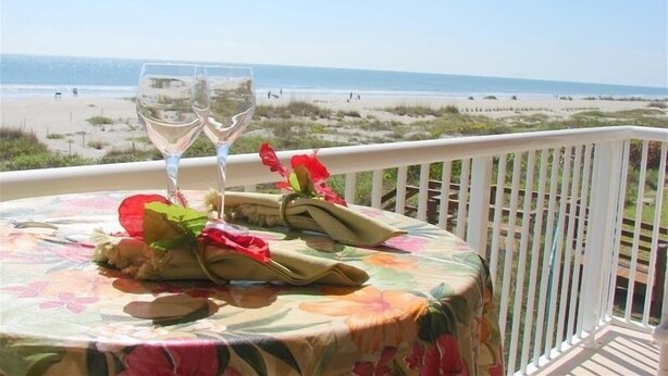 Een tafel gedekt met een tafelkleed met bloemen, twee wijnglazen en een decoratief middelpunt op een balkon met uitzicht op een strand.