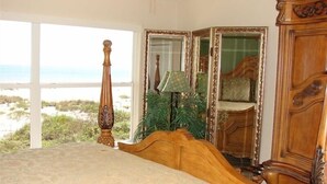 Een slaapkamer met een groot bed, een houten hoofdbord, een spiegel, een lamp en uitzicht op het strand.