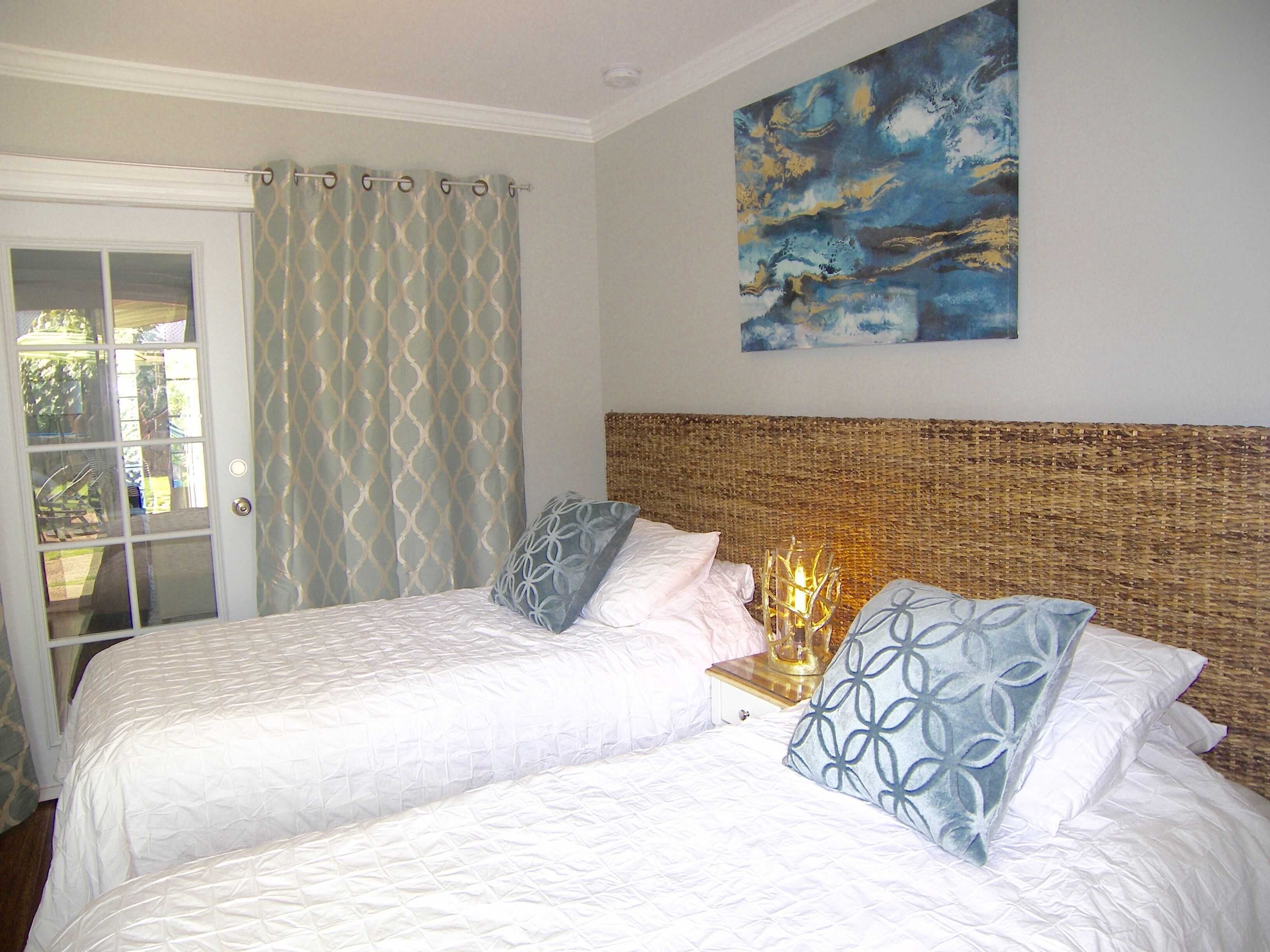 A bedroom with two beds, a wicker headboard, a window with curtains, and a painting on the wall.