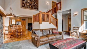 Un salon spacieux avec un canapé en cuir marron, un pouf à motifs, une table à manger en bois avec des chaises, une cheminée en pierre et un escalier en bois.