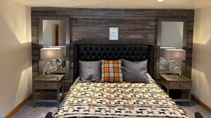Une chambre à coucher avec une tête de lit capitonnée, une couvre-lit à motifs, deux tables de chevet avec des lampes et un miroir de chaque côté.