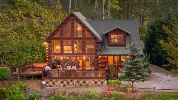 Una cabaña de troncos con terraza y zona de estar exterior, rodeada de un bosque frondoso.