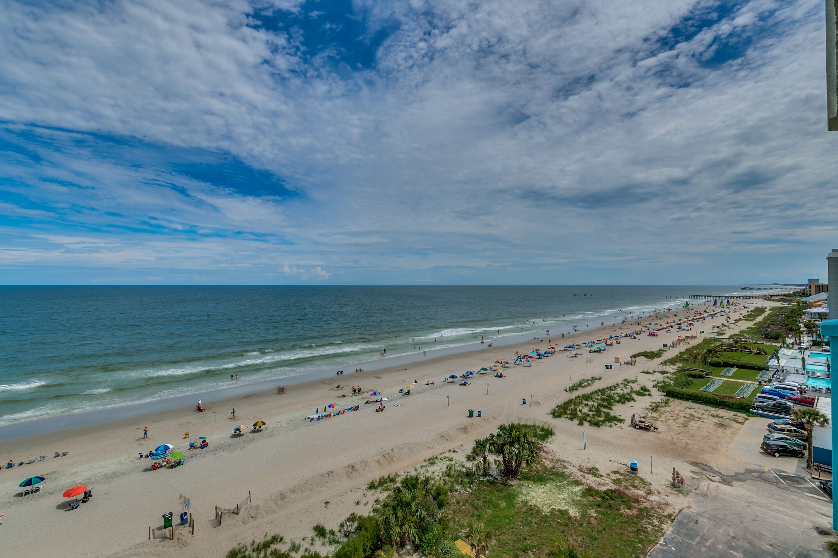 A beach with umbrellas, a pier, and a parking lot.