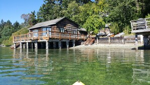 A wooden cabin with a deck extending over water, surrounded by trees. - Charming Beach Cabin Nestled in Quartermaster Harbor (Vashon)