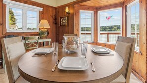 A wooden dining room with a round table set for four, a vase with seashells, and a view of a lake through large windows. - Charming Beach Cabin Nestled in Quartermaster Harbor (Vashon)