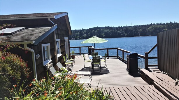 A wooden deck with outdoor seating, a green umbrella, and a view of a lake. - Charming Beach Cabin Nestled in Quartermaster Harbor (Vashon)