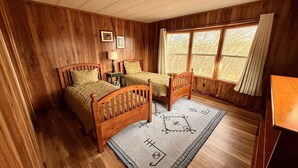 Een kamer met houten lambrisering, twee houten bedden met groene beddengoed, een kamerbreed tapijt en een raam met gordijnen.