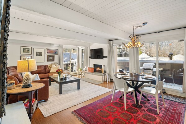 A spacious living room with a fireplace, a dining area with a chandelier, and large windows offering a view of the outdoors. - Aspen Mountain Views, Bright Corner 2BR, Pool, Hot Tub, 7-Min Walk to Gondola (Aspen)