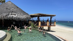 Una piscina cristalina con una estructura de techo de paja y una terraza de madera con vista a una playa.