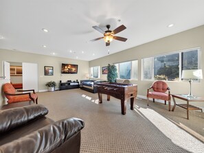 A spacious living room with a foosball table, leather sofas, a ceiling fan, and large windows.