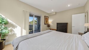 Een slaapkamer met een bed, een dressoir, een bankje, een plant in pot en een schuifdeur naar een buitenruimte.