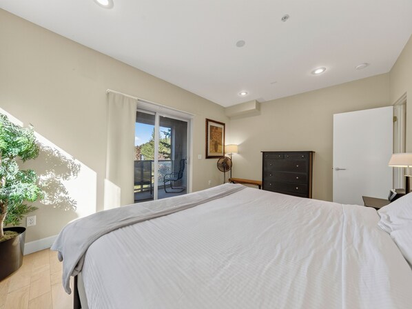 A bedroom with a bed, a dresser, a bench, a potted plant, and a sliding glass door leading to an outdoor area.