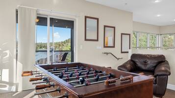 A room with a foosball table, a leather sofa, and a view of a forest through large windows.