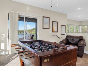 A room with a foosball table, a leather sofa, and a view of a forest through large windows.