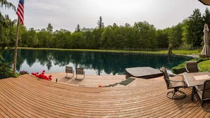 Una terraza de madera con mobiliario exterior, una bandera y vistas a un lago rodeado de árboles.