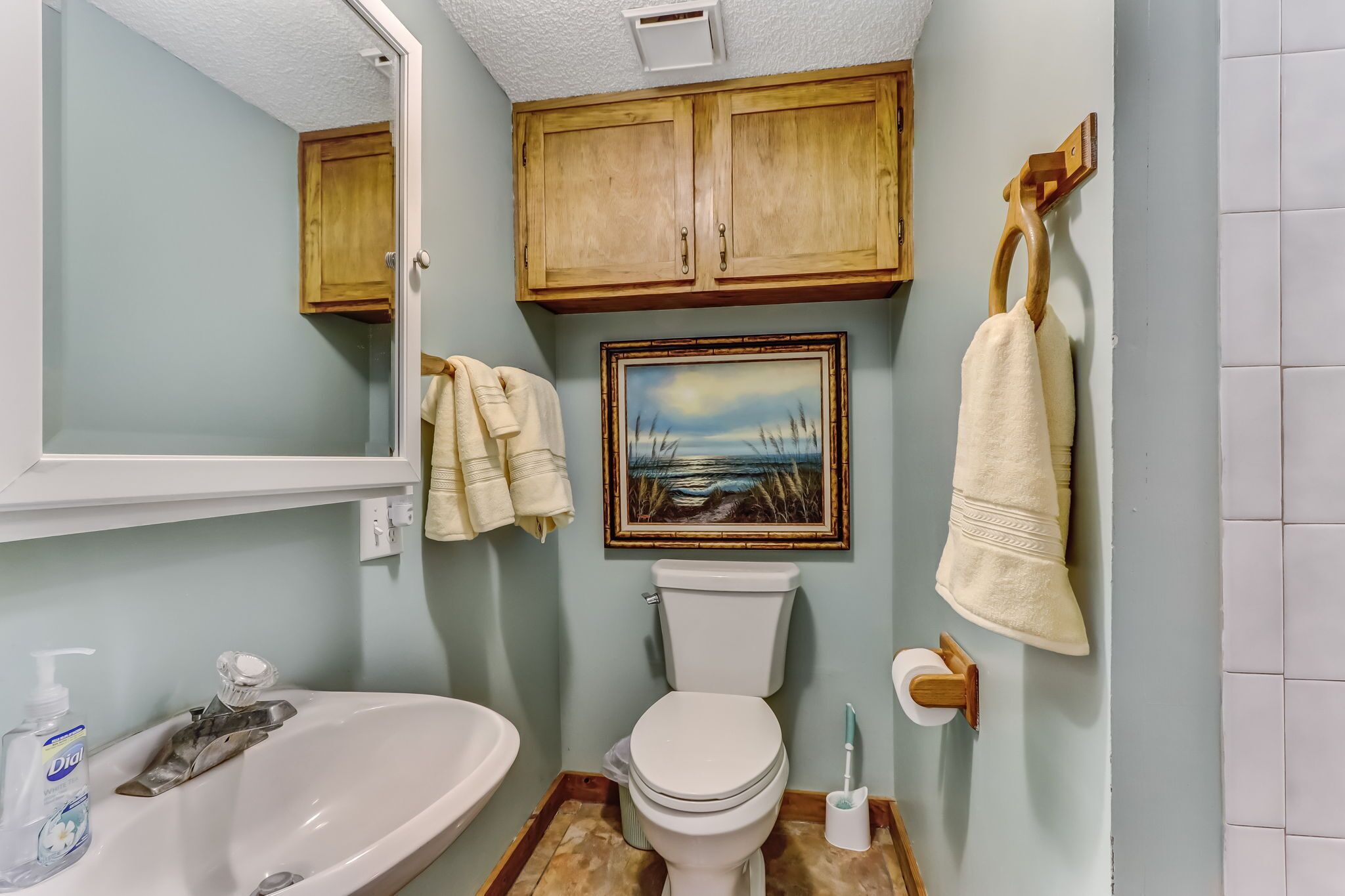 A bathroom with a sink, toilet, wooden cabinets, a framed picture, and hanging towels.