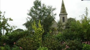 A garden with a white plastic table and chairs, dense greenery, and a church steeple in the background.