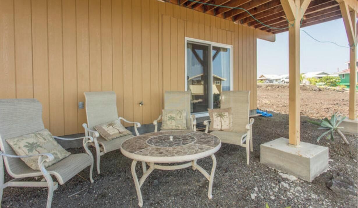 Een terras met wicker meubels en een ronde tafel, naast een houten gebouw met een glazen deur.