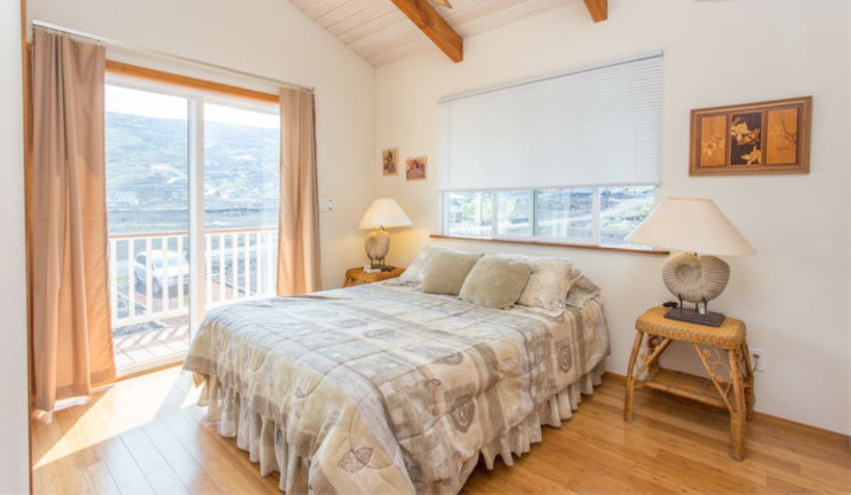 A bedroom with a large bed, a ceiling fan, a window with a view, and a small table with a lamp.