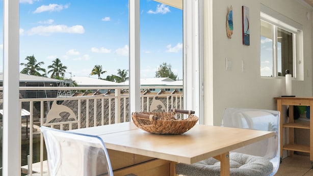 A modern dining area with a wooden table and chairs, a large window with a view of palm trees, and a balcony with a railing. - Nov 18-Jan 1 Open... Live Like a Local*Waterview*Pet Friendly*Key Largo MM 99.5*Manatees (Key Largo)