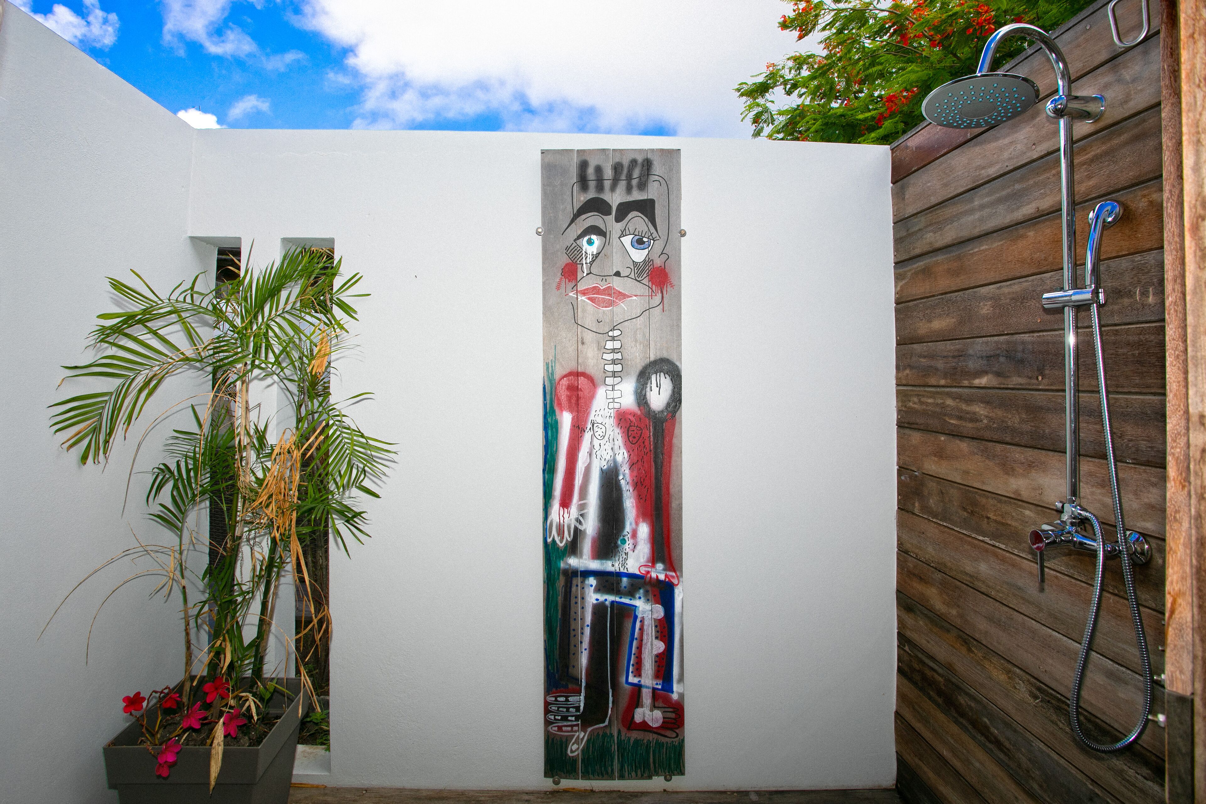 Un baño con cabina de ducha de madera, pared blanca con un mural vertical y una planta en maceta con flores rojas.