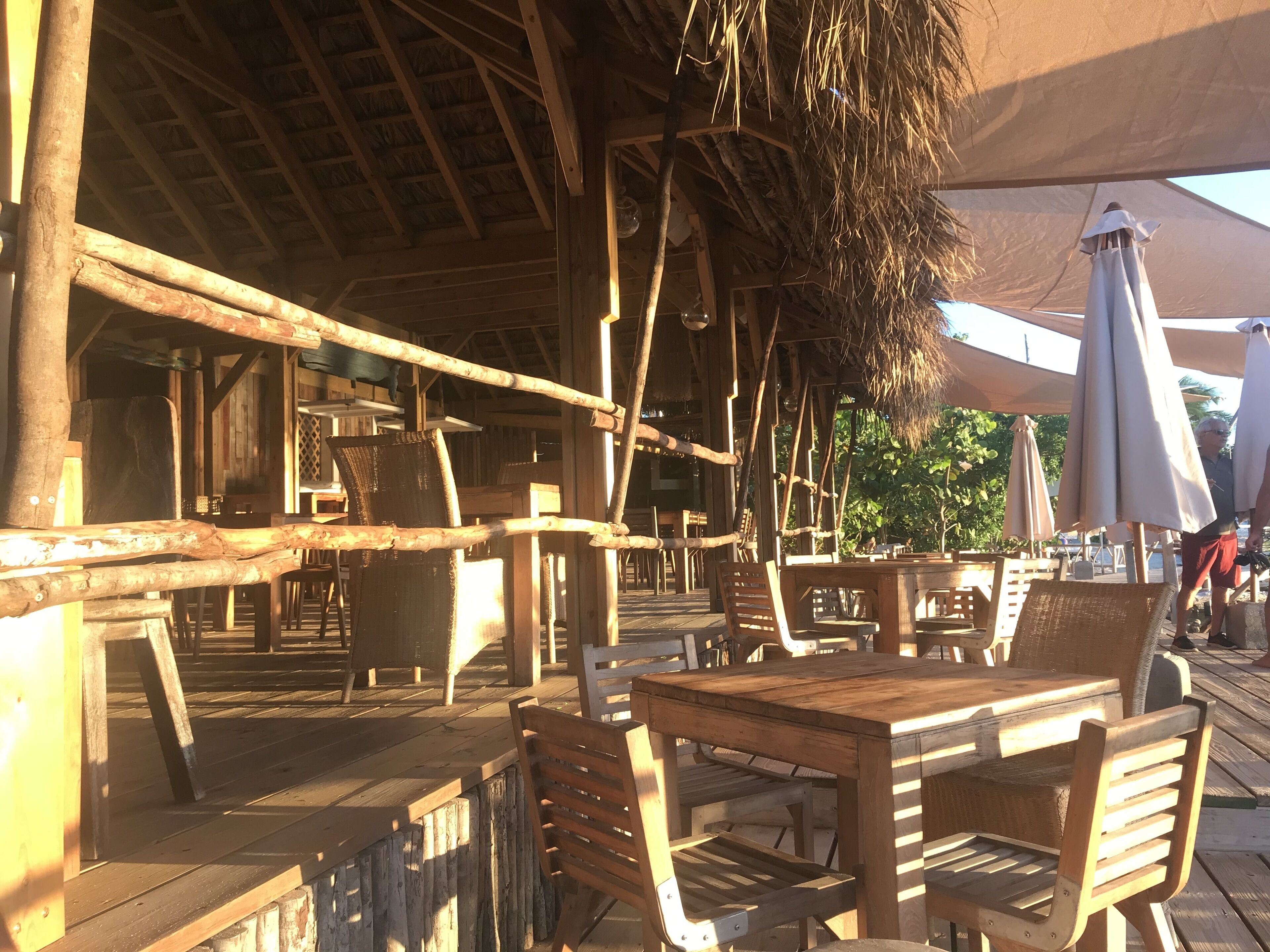 Eine hölzerne Terrasse im Freien mit Tischen, Stühlen, einem Reetdach und Sonnenschirmen.