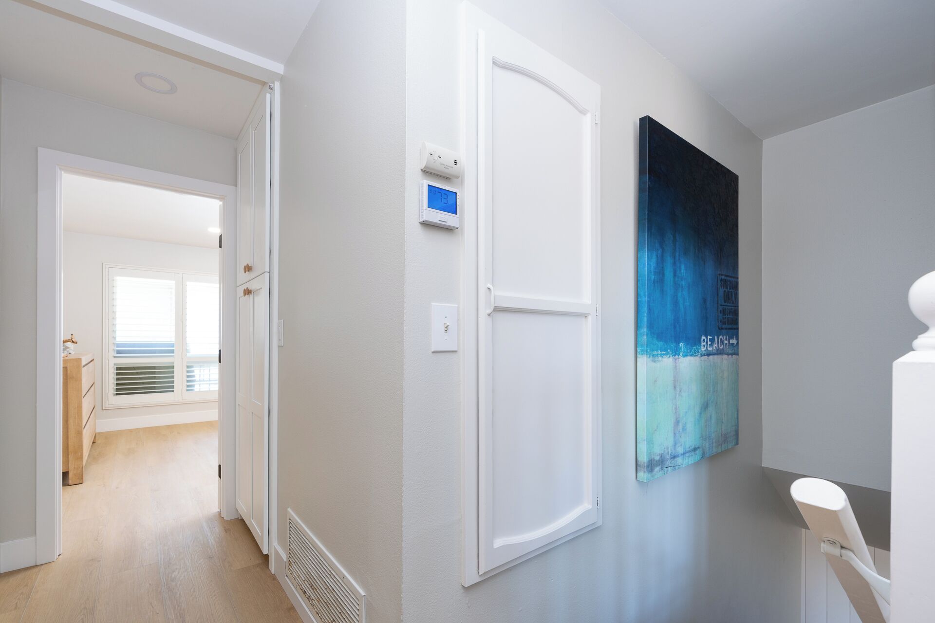 A hallway with a white door, a thermostat, and a wall-mounted artwork depicting a beach scene.