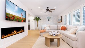 Un salon moderne doté d’une cheminée, d’un grand téléviseur à écran plat, d’un ventilateur de plafond et d’une grande fenêtre avec des stores.