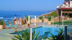 Un espace piscine agrémenté de chaises longues et de plantes en pot, offrant une vue sur une plage et l’océan.