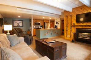 A living room with a sofa, a coffee table, a fireplace, and a kitchen area with wooden cabinets and a TV mounted on the wall.